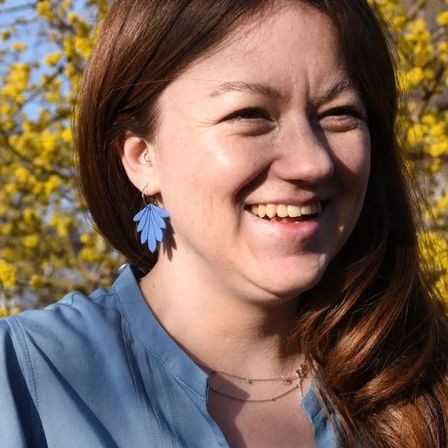 Eine Frau mit blauem Hemd und blauen Ohrringen lächelt vor einem gelben Baum.