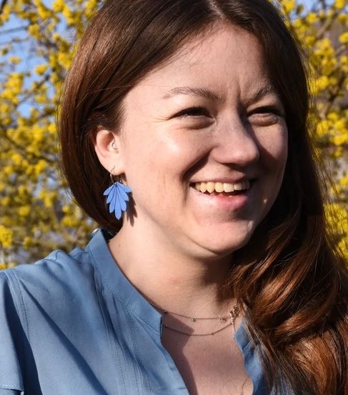 Eine Frau mit blauem Hemd und blauen Ohrringen lächelt vor einem gelben Baum.