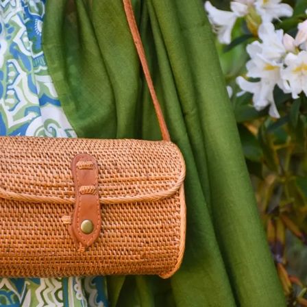 Eine Person, die einen grünen Schal trägt, trägt eine braune Korbtasche mit einem Lederriemen und einem braunen Knopf. Dahinter ist eine weiße Blume mit grünen Blättern zu sehen.