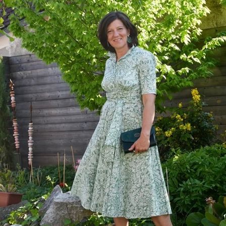 Eine Frau in einem Blumenkleid steht lächelnd im Garten und hält eine schwarze Handtasche in der Hand. Dahinter steht ein Holzzaun und ein Baum mit gelben Blumen.