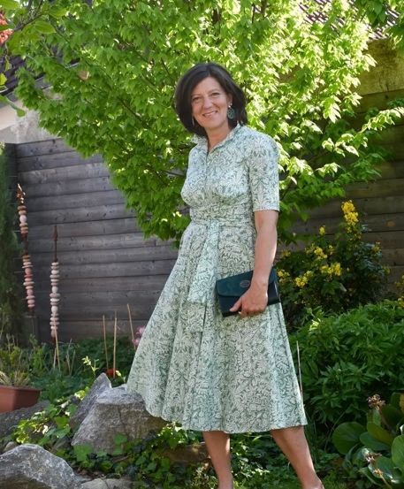 Eine Frau in einem Blumenkleid steht lächelnd im Garten und hält eine schwarze Handtasche in der Hand. Dahinter steht ein Holzzaun und ein Baum mit gelben Blumen.