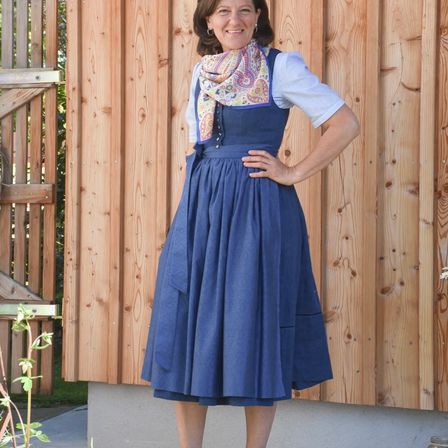 Eine Frau steht lächelnd vor einer Holzwand. Sie trägt ein blaues Kleid mit weißem Schürzen und ein gemustertes Tuch.