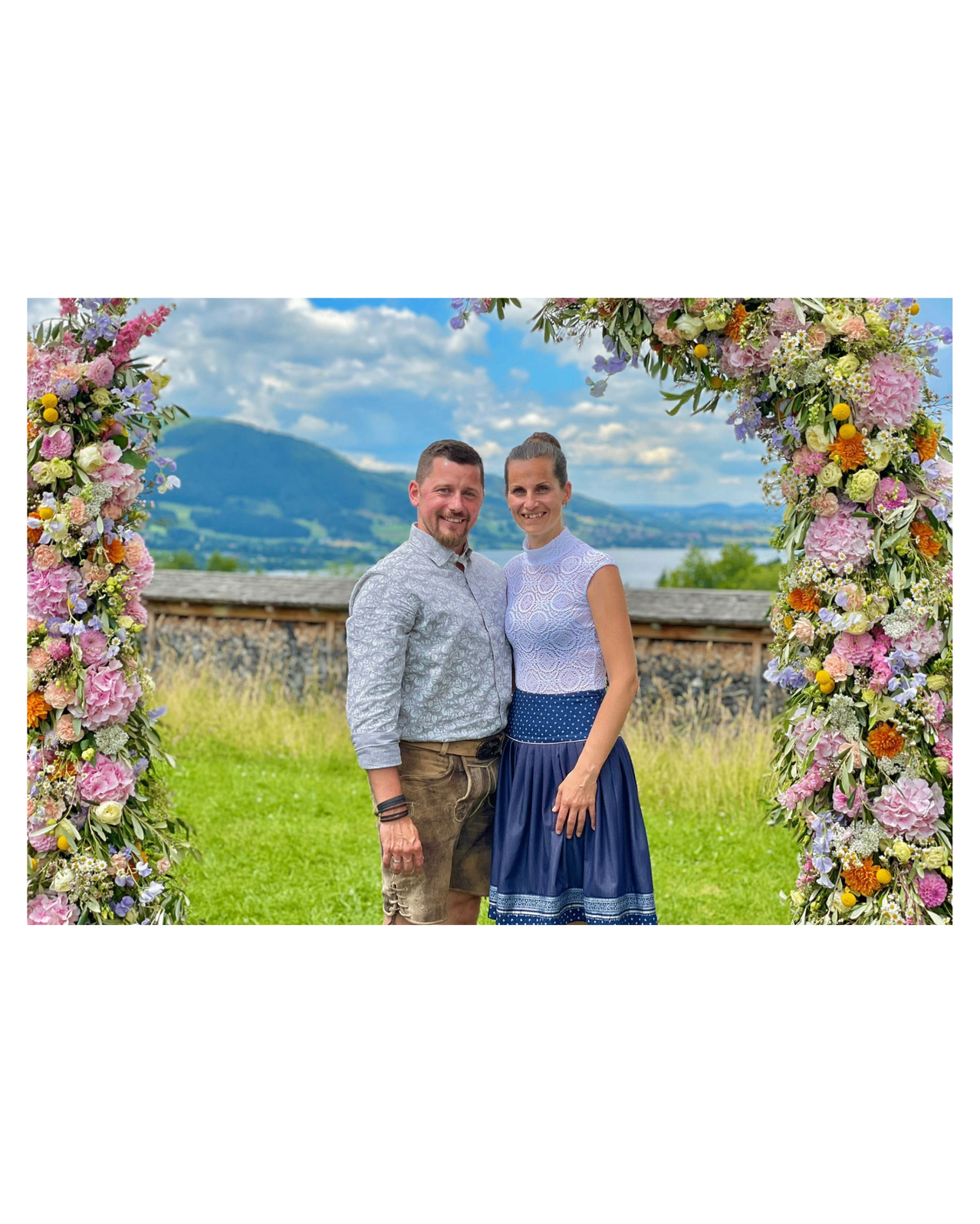 Ein lächelndes Paar steht unter einem Blumenbogen an einem sonnigen Tag mit Bergen und einem See im Hintergrund.