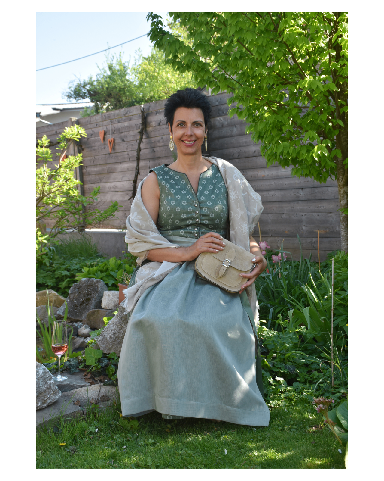 Eine Frau im Garten in einem grünen Kleid und einem Umhang, hält eine beige Tasche und lächelt, mit einem Glas Wein in der Nähe.