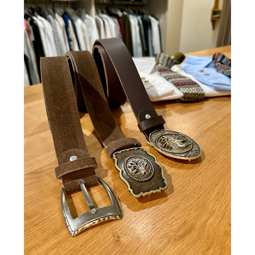 Zwei braune Ledergürtel mit verzierten Schnallen liegen auf einem Holztisch, mit gefalteter Kleidung im Hintergrund.