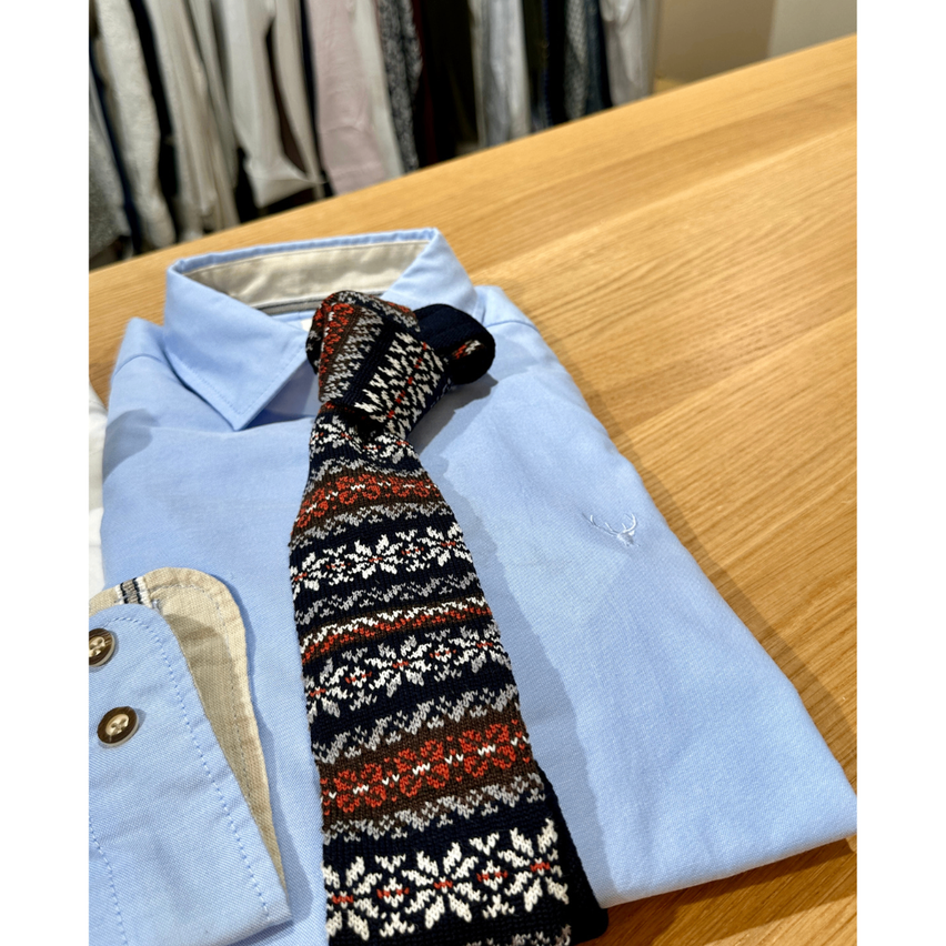Ein hellblaues Hemd mit einer gestrickten Krawatte auf einem Holztisch. Im Hintergrund sind weitere Hemden zu sehen.