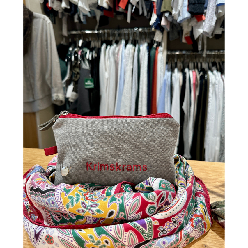 Ein grauer Krimskrams-Beutel auf einem bunten Schal mit Kleidung im Hintergrund.