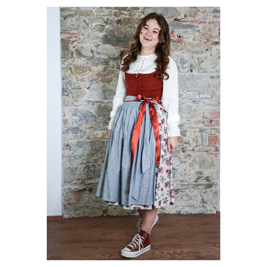 Eine Frau in traditioneller deutscher Kleidung steht vor einer Ziegelwand, lächelt und trägt ein rot-weißes Kleid mit einem Blumenrock.