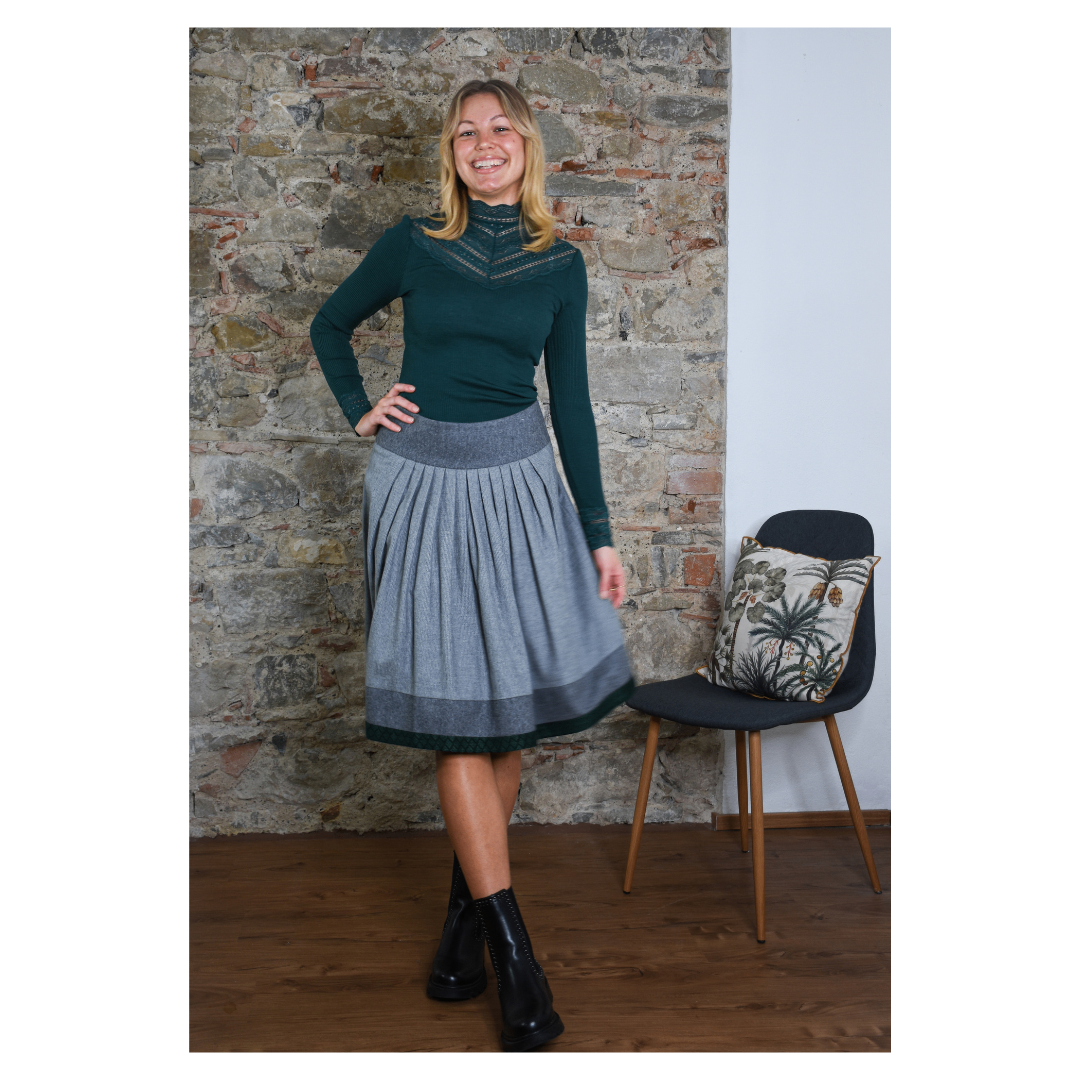 Eine Frau in einem grünen Langarmshirt und einem grauen Rock steht lächelnd vor einer Backsteinmauer. Sie trägt schwarze Stiefel. Ein Stuhl mit Kissen steht in der Nähe.