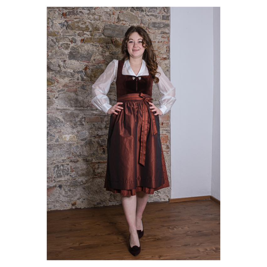 Eine Frau steht drinnen, trägt ein braunes Kleid und eine weiße Bluse, mit den Händen auf den Hüften. Sie lächelt und posiert für ein Foto.