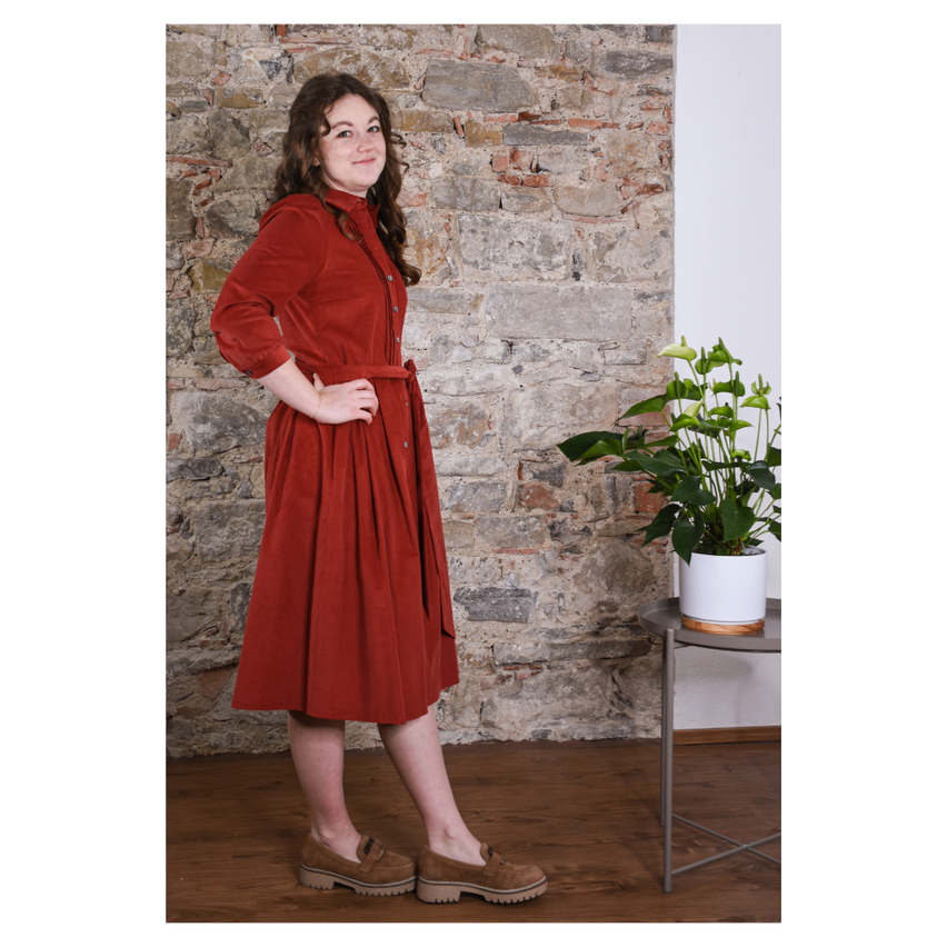 Eine Frau steht mit den Händen auf den Hüften, trägt ein rotes Samtkleid und braune Stiefel. Sie steht vor einer Ziegelmauer und neben einem Topf mit einer Pflanze auf einem runden Tisch.