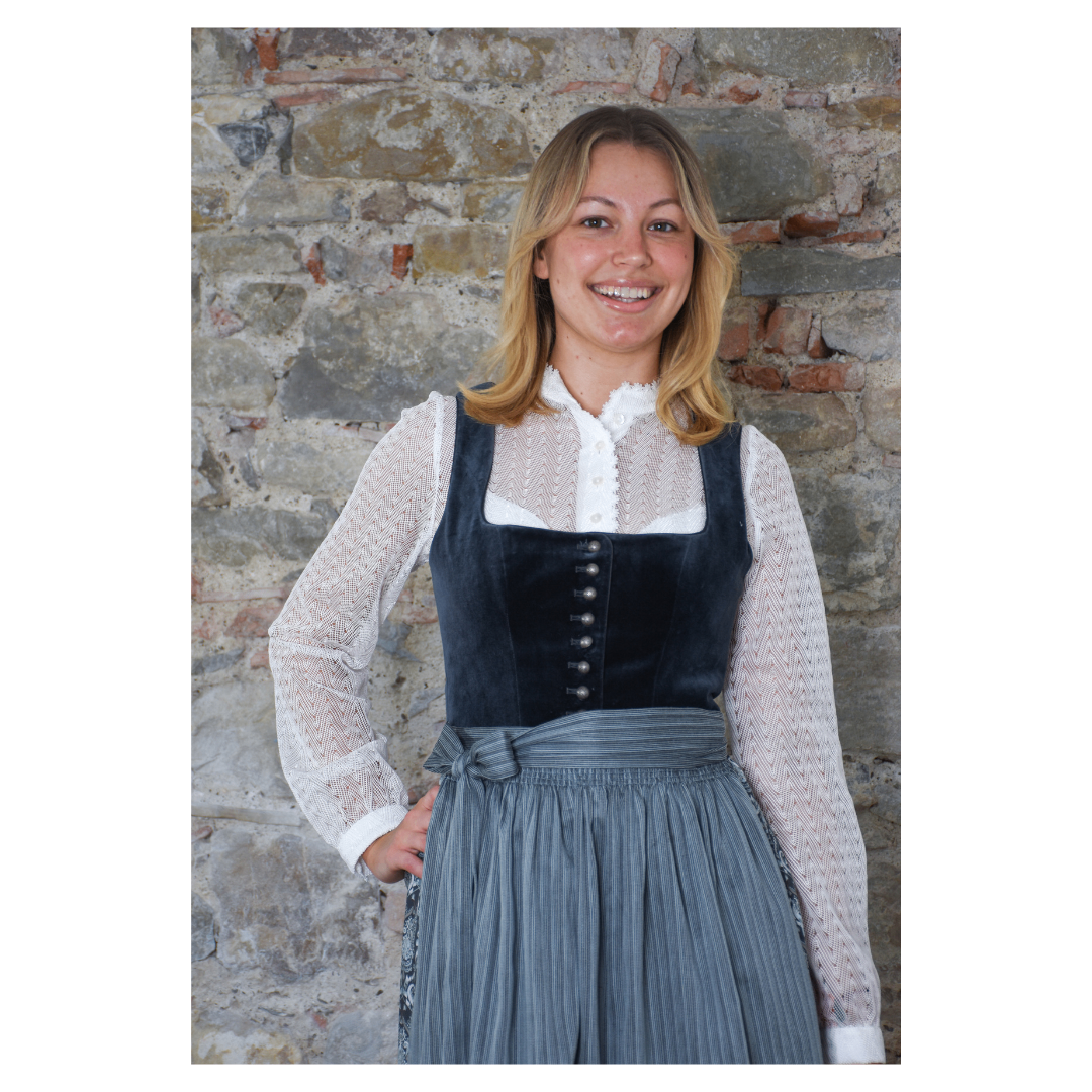 Eine Frau in traditioneller Kleidung steht lächelnd vor einer Ziegelmauer. Sie trägt ein weißes Oberteil mit einem schwarzen Westchen und einem plissierten Rock.