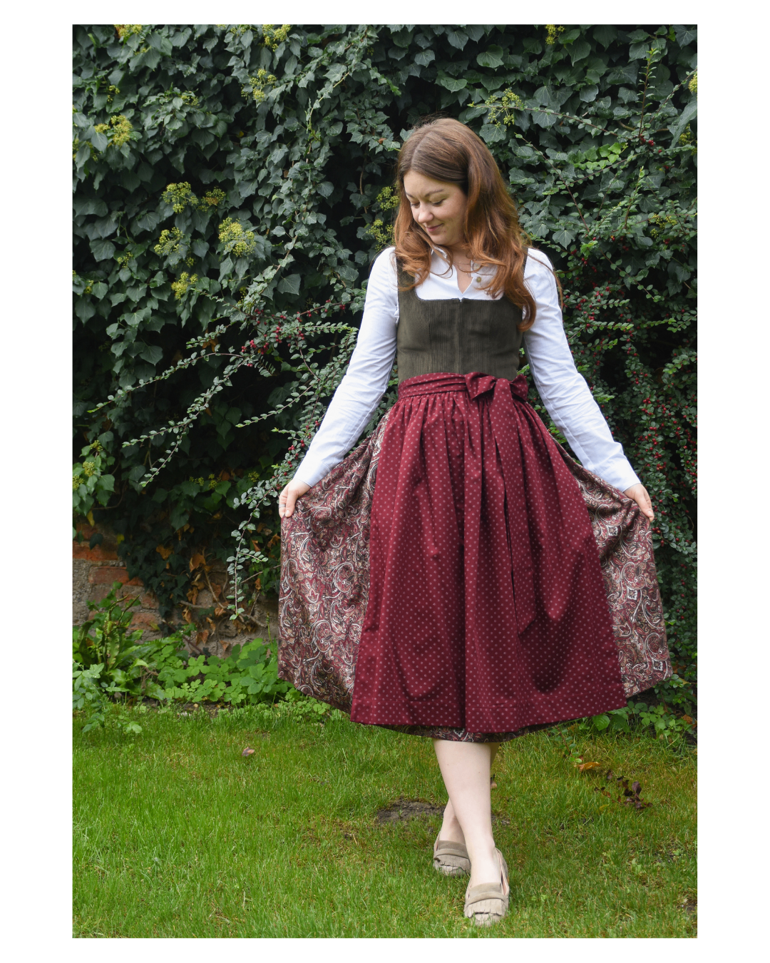 A woman in a maroon polka dot skirt with a brown vest stands in front of green foliage.