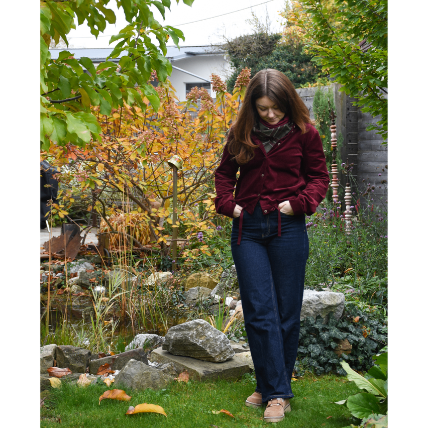 A woman stands in a garden with autumn leaves, wearing a maroon sweater and blue jeans, looking down at her hands.