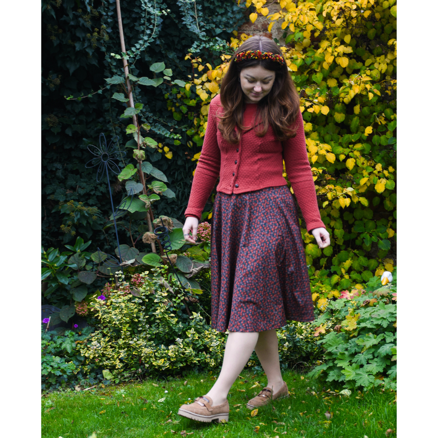 A woman wearing a red cardigan and floral skirt walks in a garden with yellow and green foliage. She has a flower crown on her head.