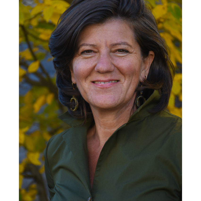 A woman with short dark hair smiles for the camera against a backdrop of yellow leaves. She wears a green shirt and earrings.