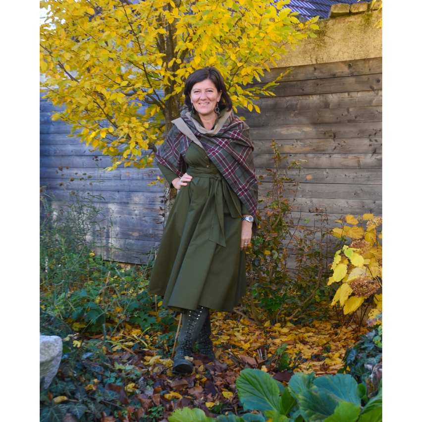 A woman in a green dress and plaid scarf stands in a garden, smiling. She is surrounded by yellow leaves and a tree with autumn foliage. Behind her, a wooden wall is visible.