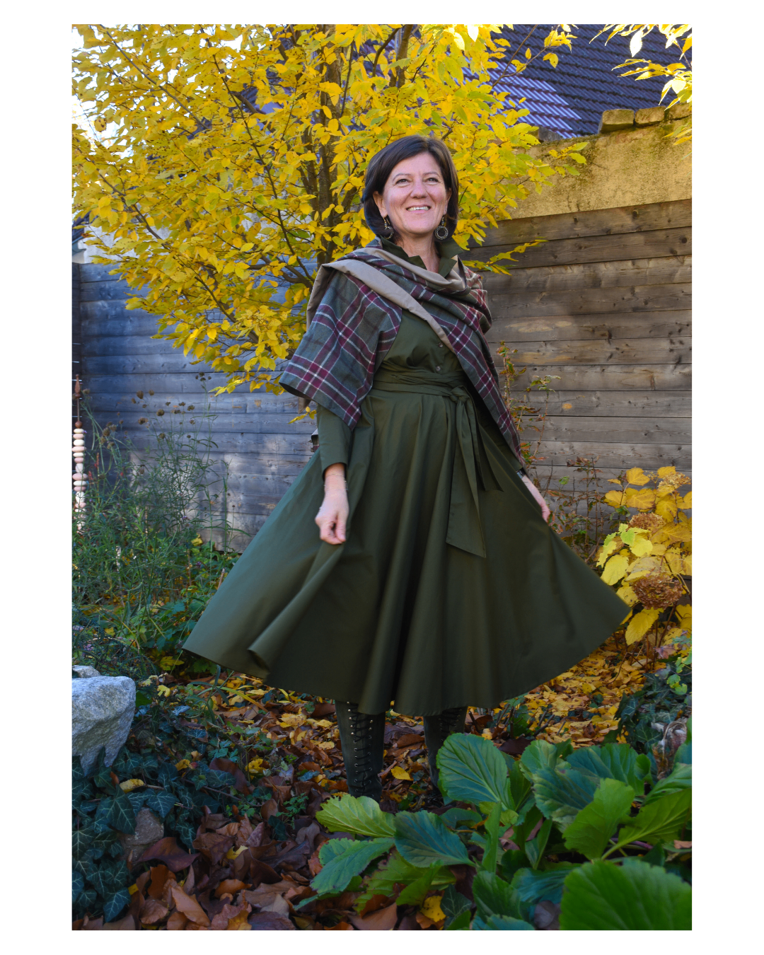 A woman stands outdoors in a garden, smiling, wearing a green dress with a plaid scarf around her neck.