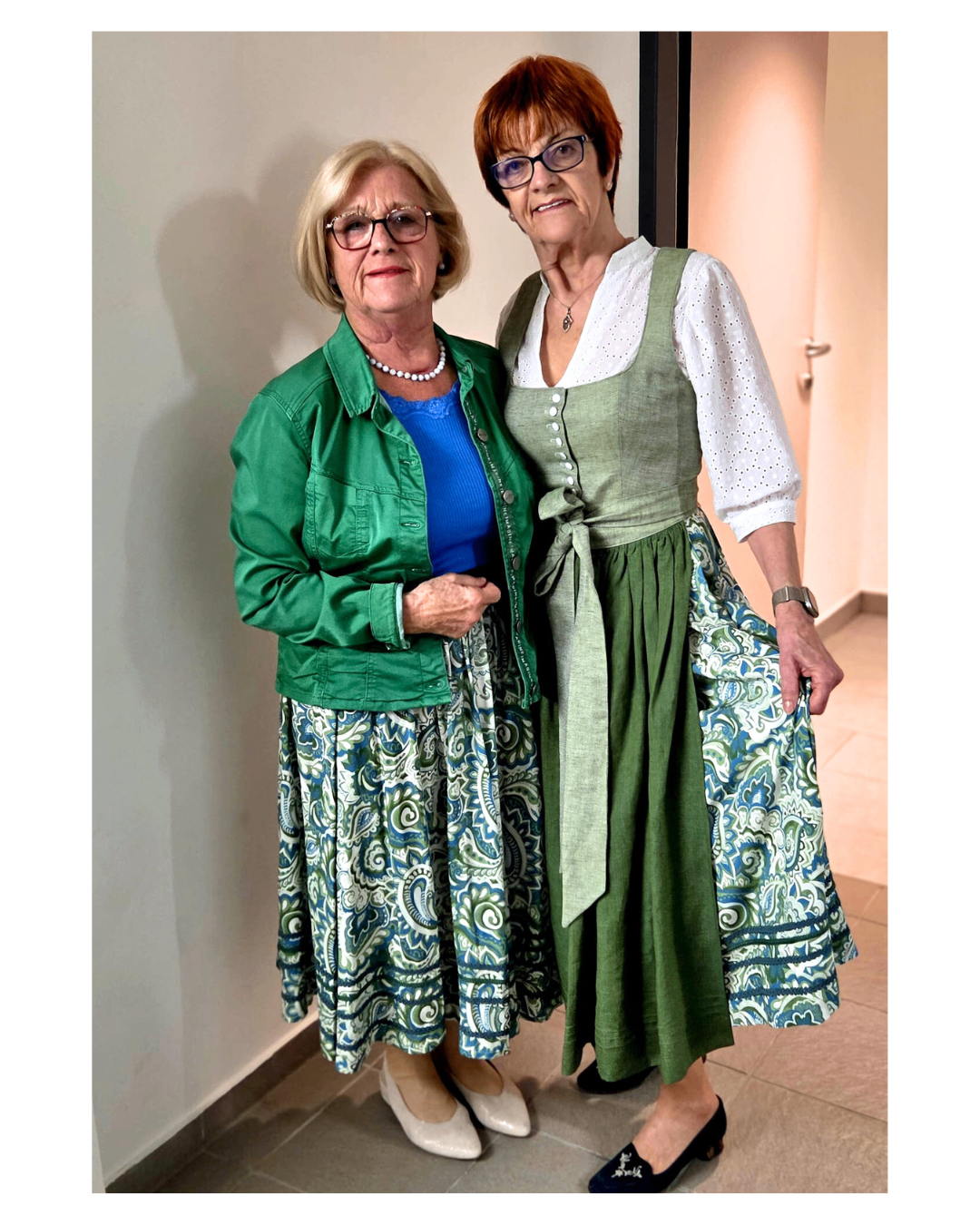 Zwei ältere Frauen stehen nebeneinander und lächeln. Die Frau auf der linken Seite trägt eine grüne Jacke und ein blaues Oberteil. Die Frau auf der rechten Seite trägt ein traditionelles grünes Kleid mit einem weißen Oberteil.