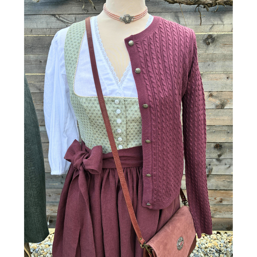 Ein Mannequin zeigt einen karmesinroten Cardigan mit Knöpfen über einem weißen Blusenoberteil, einem grünen Schürze und einem burgunderroten Rock mit einer Schleife, zusammen mit einer braunen Ledertasche.