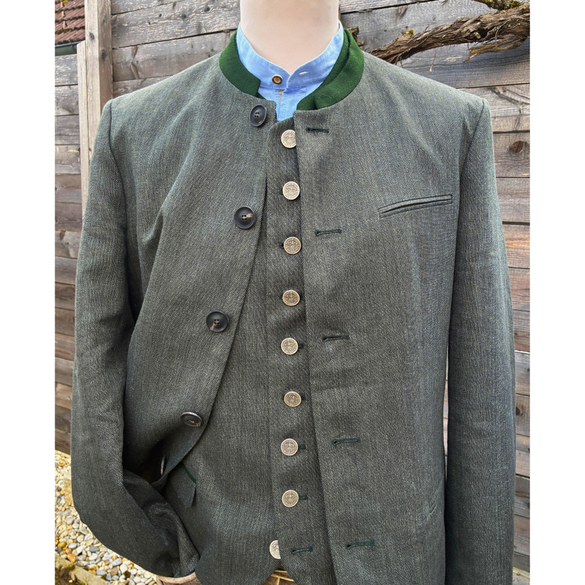 Ein grüner Anzug mit goldenen Knöpfen und einem grünen Kragen wird auf einem hölzernen Hintergrund ausgestellt. Der Anzug hat ein blaues Hemd darunter und mehrere Taschen.