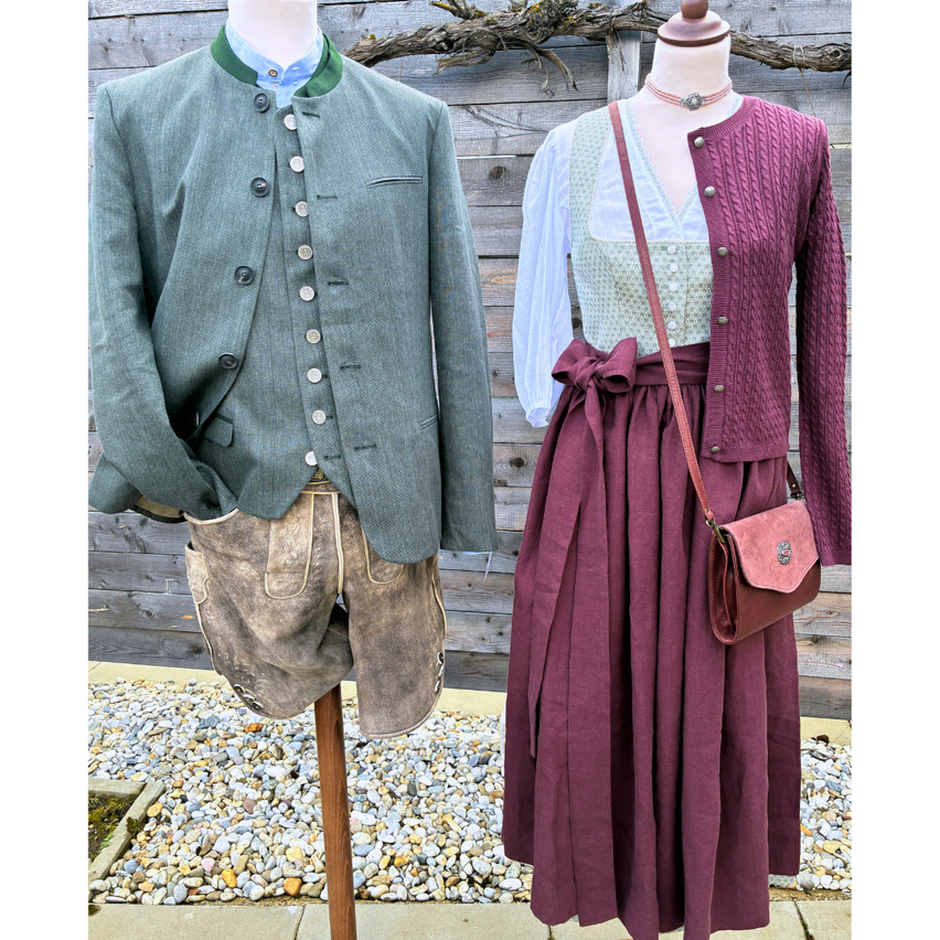 Zwei Mannequins präsentieren traditionelle Kleidung; eines trägt eine grüne Jacke mit Knöpfen und braune Shorts, das andere einen braunen Rock und eine gestrickte Strickjacke mit einer braunen Handtasche.