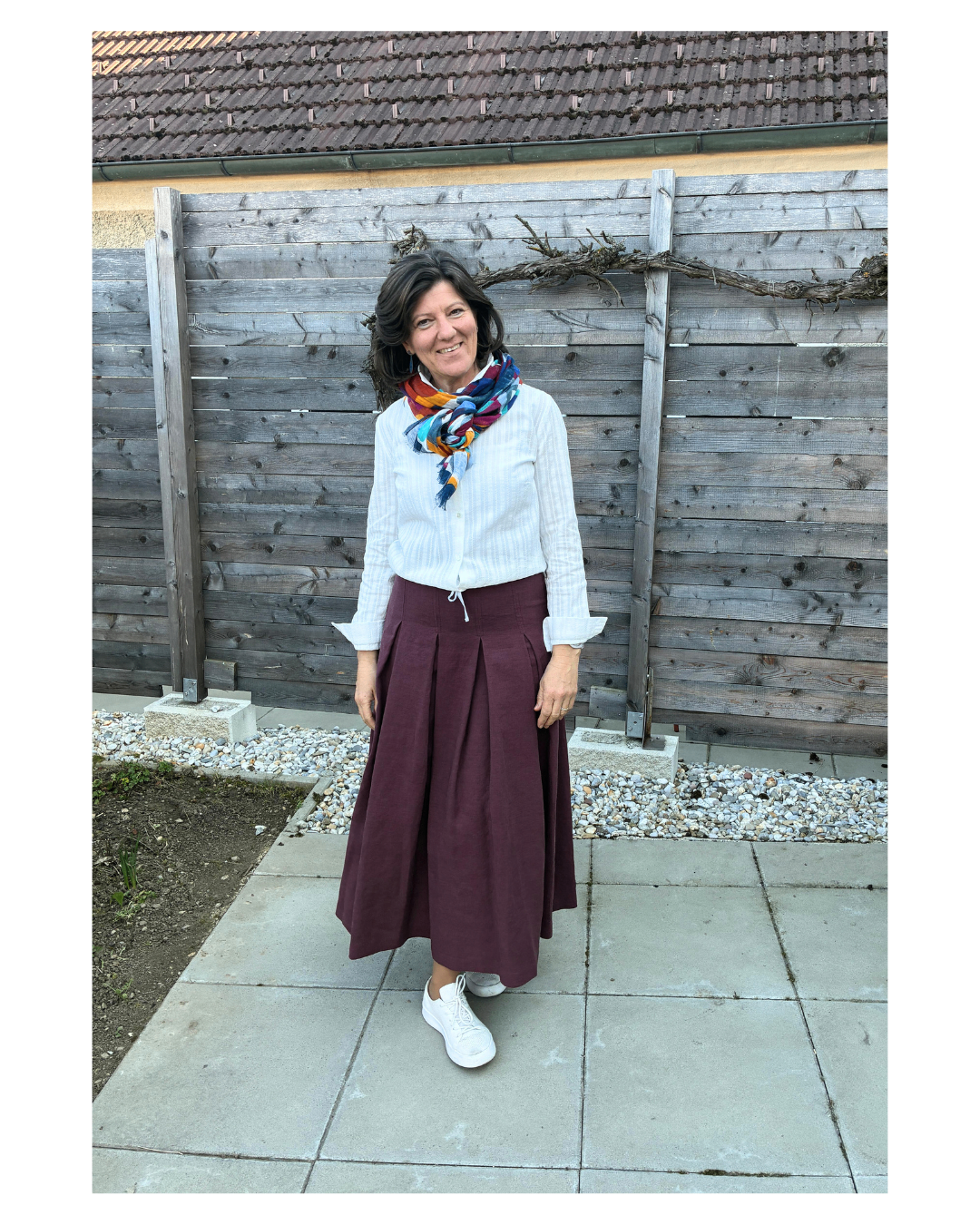 Eine Frau in einem weißen Oberteil, einer Weinroten Rock und einem bunten Schal steht lächelnd vor einem Holzzaun.