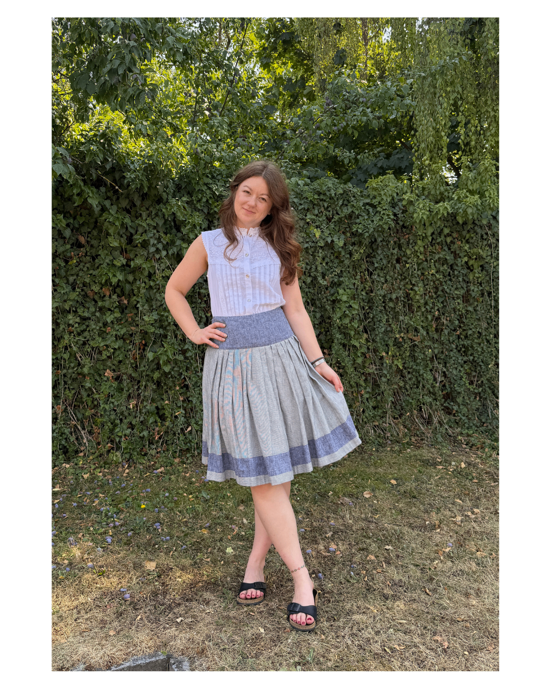 Eine Frau steht in einem Garten, lächelt und trägt eine weiße Bluse und einen grauen Rock mit blauem Streifen. Sie ist barfuß und hat die Hände auf den Hüften.