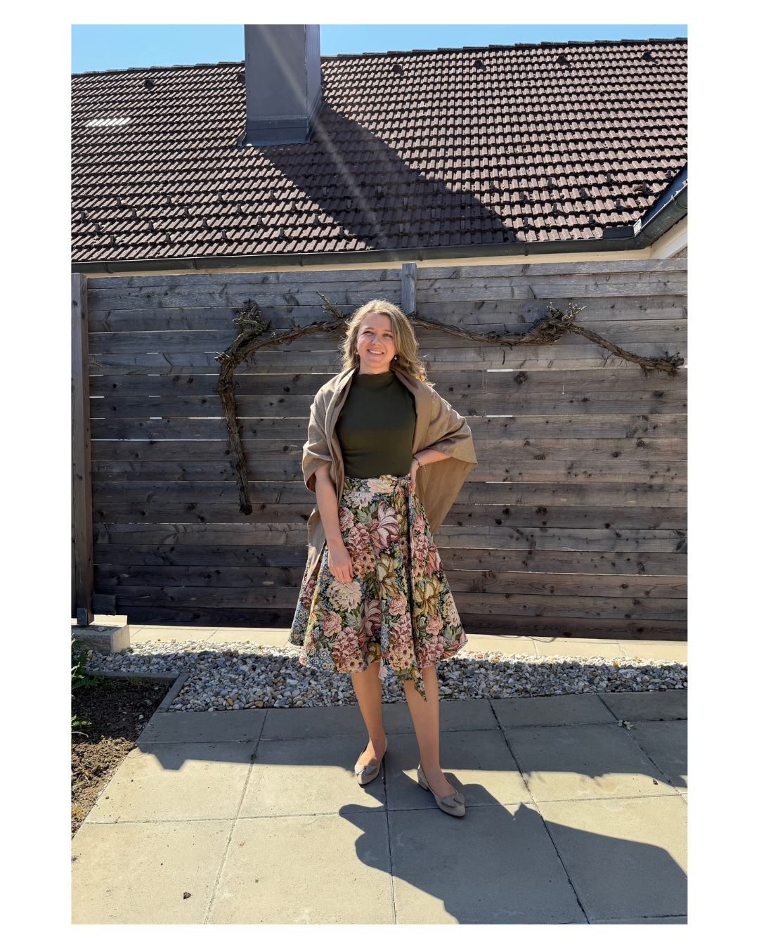 Eine Frau mit blondem Haar steht vor einem Holzzaun, trägt einen Blumenrock und ein grünes Oberteil und lächelt für ein Foto.