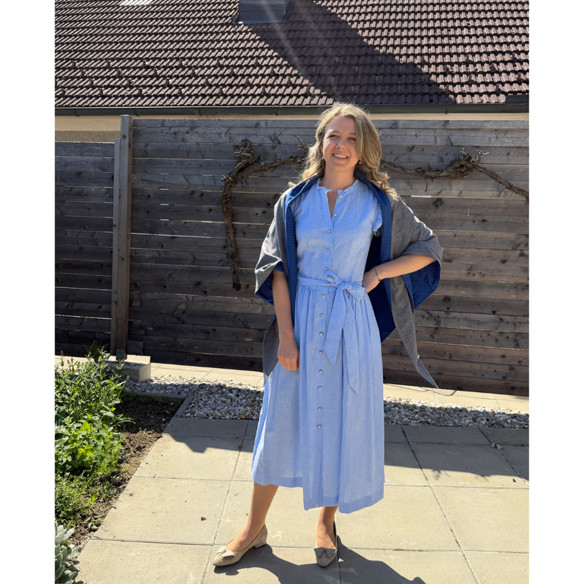 Eine Frau in einem blauen Kleid und einem grauen Schal steht vor einem Holzzaun mit einem Haus und Pflanzen im Hintergrund.