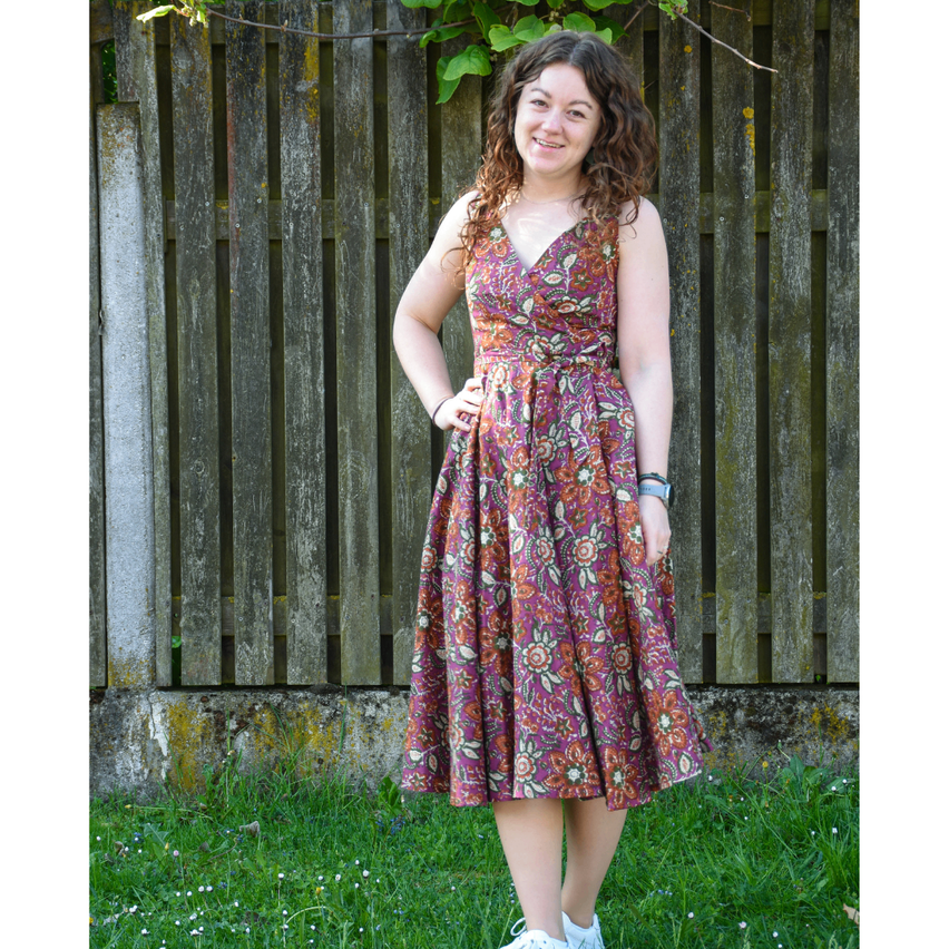 Eine Frau in einem Blumenkleid steht vor einem Holzzaun. Sie lächelt und trägt eine Uhr.
