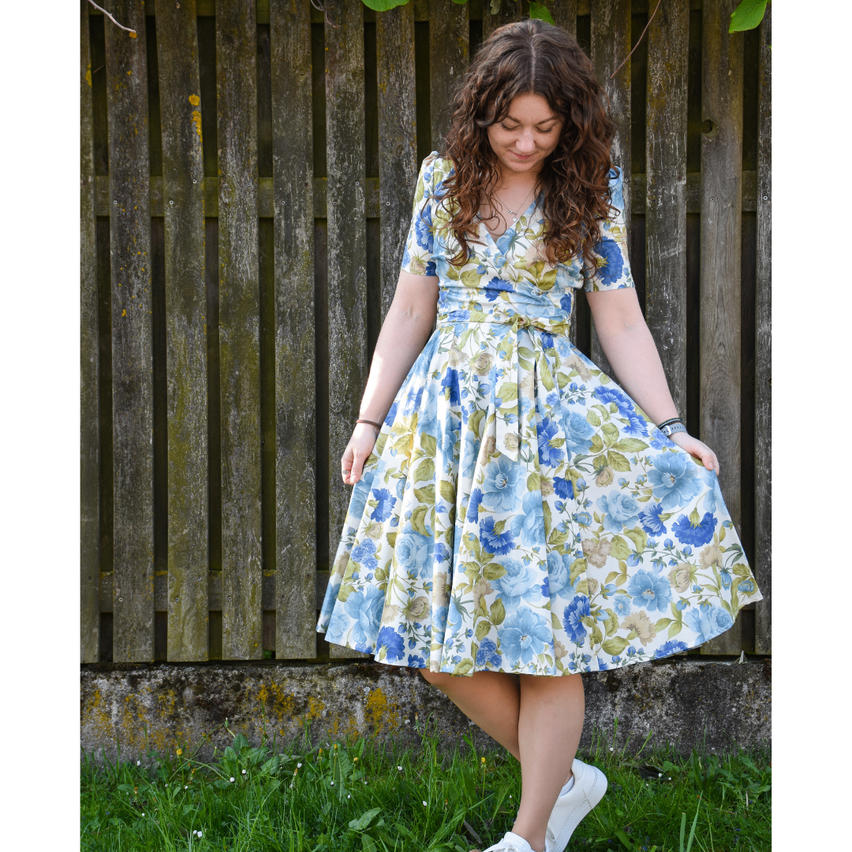 Eine Frau in einem Blumenkleid steht vor einem Holzzaun, lächelt und posiert für ein Foto.