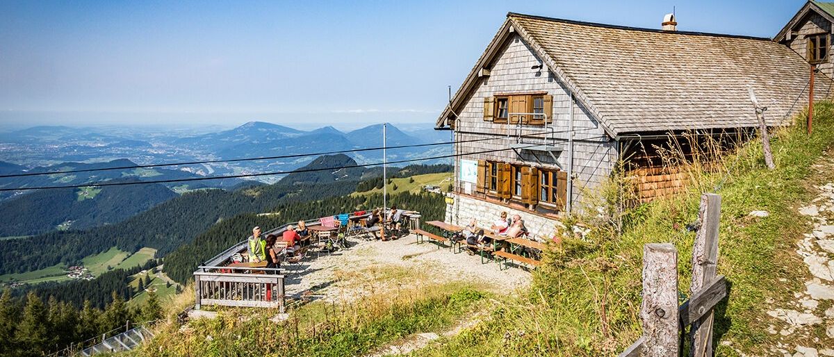 Eine rustikale Berghütte mit Bänken und Tischen steht auf einem Hügel. Die Leute entspannen sich auf den Bänken und genießen die Aussicht auf die umliegenden Berge.