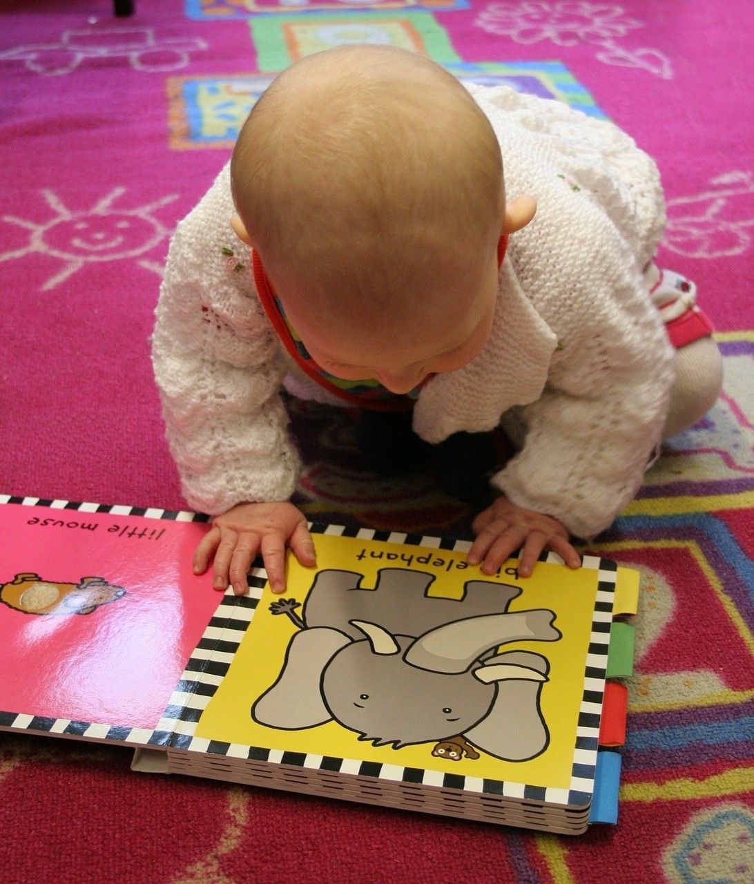 Ein Baby kriecht auf einem rosa Teppich und hält ein offenes Buch mit einer Elefanten-Illustration auf dem Cover. Das Buch hat eine gelbe Seite und einen schwarz-weiß gestreiften Rand.