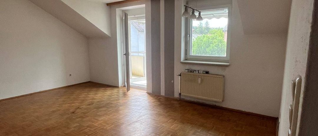 Ein leeres Dachzimmer mit Holzböden, weißen Wänden und einem Fenster mit Blick auf Bäume draußen.