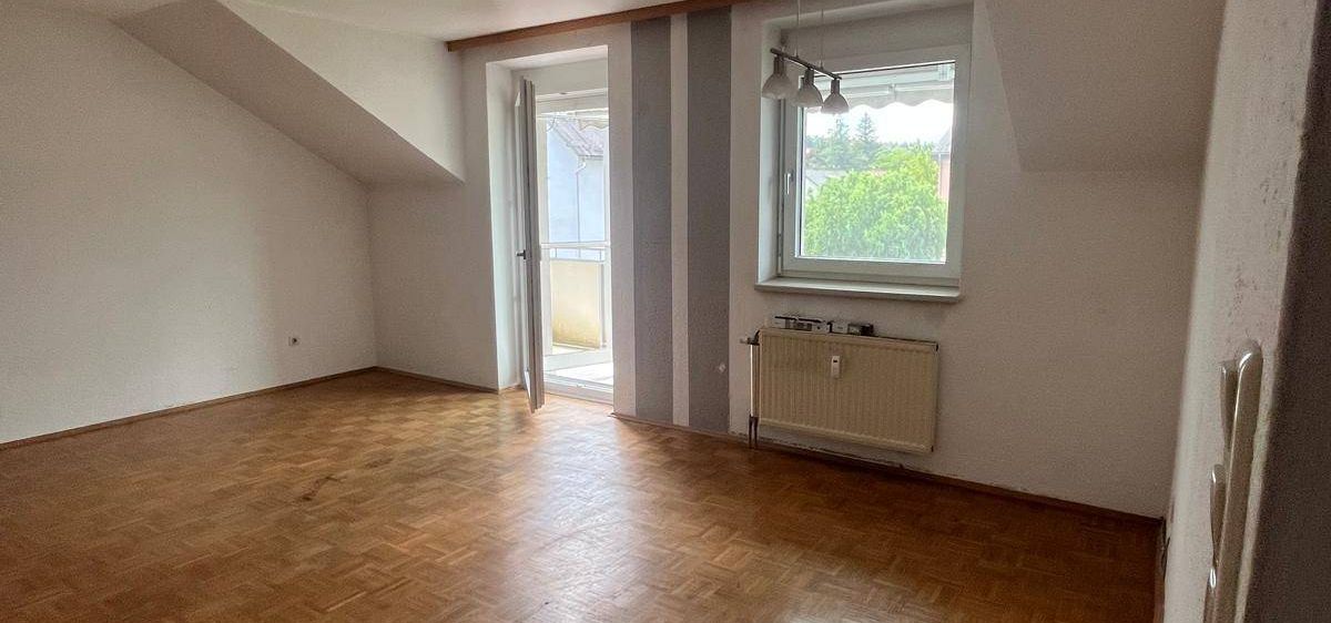 Ein leeres Dachzimmer mit Holzböden, weißen Wänden und einem Fenster mit Blick auf Bäume draußen.