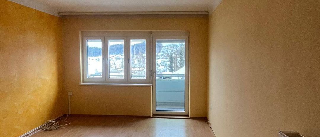 Ein leerer Raum mit Holzböden, gelben Wänden und einer Schiebetür aus Glas, die zu einem Balkon führt.