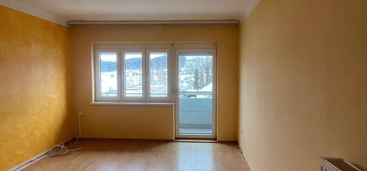Ein leerer Raum mit Holzböden, gelben Wänden und einer Schiebetür aus Glas, die zu einem Balkon führt.