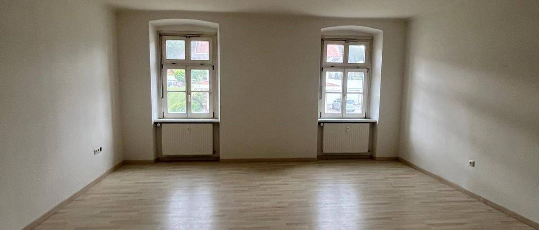 Ein leerer Raum mit weißen Wänden und Holzböden. Zwei Fenster sind sichtbar, mit weißen Rahmen und einem kleinen Heizkörper darunter. Der Raum wird durch Sonnenlicht beleuchtet, das durch die Fenster scheint.