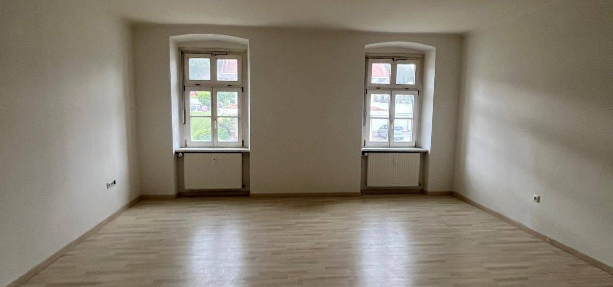 Ein leerer Raum mit weißen Wänden und Holzböden. Zwei Fenster sind sichtbar, mit weißen Rahmen und einem kleinen Heizkörper darunter. Der Raum wird durch Sonnenlicht beleuchtet, das durch die Fenster scheint.