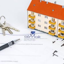 Ein kleines Hausmodell mit Schlüsseln und einem Stift auf einem Vertragspapier. Das Haus ist orange und gelb. Es hat ein Logo auf der Vorderseite.