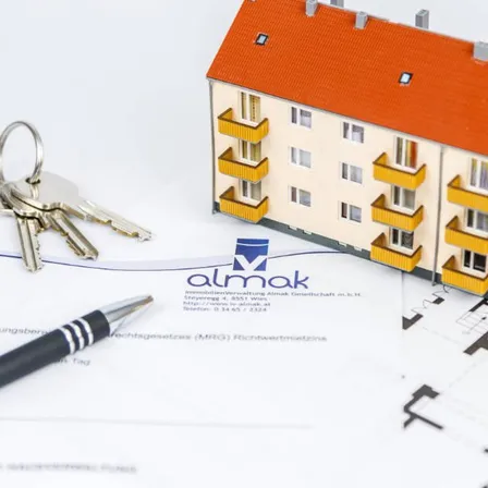 Ein kleines Hausmodell mit Schlüsseln und einem Stift auf einem Vertragspapier. Das Haus ist orange und gelb. Es hat ein Logo auf der Vorderseite.