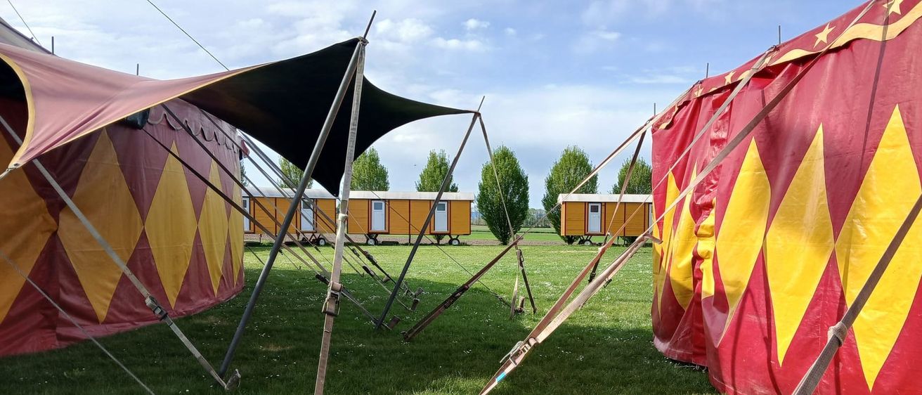 Ein Zirkuszelt ist teilweise entlüftet auf einem Grasfeld. In der Nähe sind mehrere kleine orangefarbene Wohnwagen in einer Reihe geparkt. Der Himmel ist blau mit einigen Wolken.