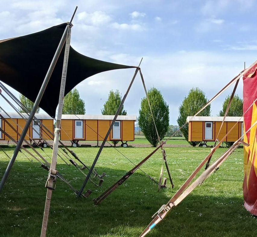 Ein Zirkuszelt ist teilweise entlüftet auf einem Grasfeld. In der Nähe sind mehrere kleine orangefarbene Wohnwagen in einer Reihe geparkt. Der Himmel ist blau mit einigen Wolken.