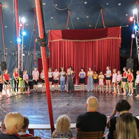 Eine Gruppe von Kindern steht auf einer Zirkusbühne unter einem roten Vorhang mit Sternen und Scheinwerfern. Zuschauer schauen vom Publikum aus zu.