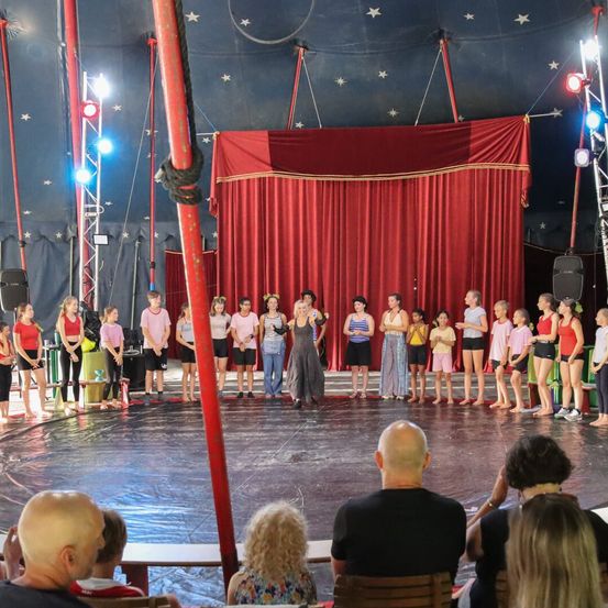 Eine Gruppe von Kindern steht auf einer Zirkusbühne unter einem roten Vorhang mit Sternen und Scheinwerfern. Zuschauer schauen vom Publikum aus zu.