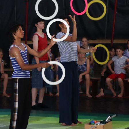 Zwei junge Darsteller jonglieren mit mehreren farbigen Ringen, während mehrere andere Kinder sie von den Seitenlinien aus beobachten.