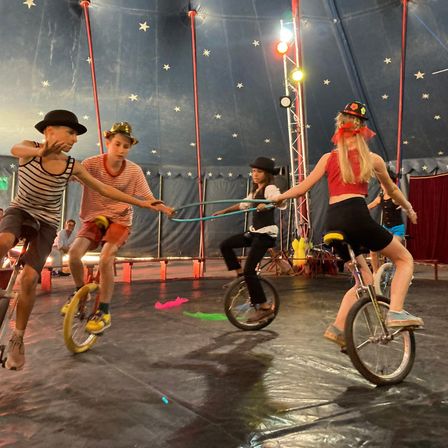 Eine Gruppe von Kindern fährt auf Einrädern unter einem Zirkuszelt. Eine Frau hält eine Schnur zum Ausgleich.