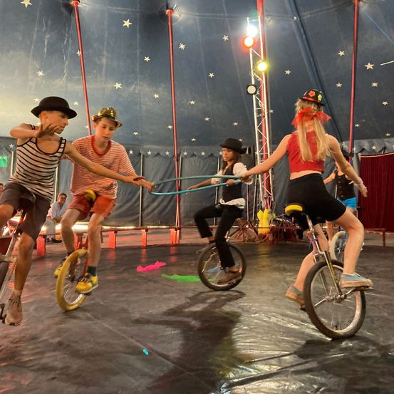 Eine Gruppe von Kindern fährt auf Einrädern unter einem Zirkuszelt. Eine Frau hält eine Schnur zum Ausgleich.