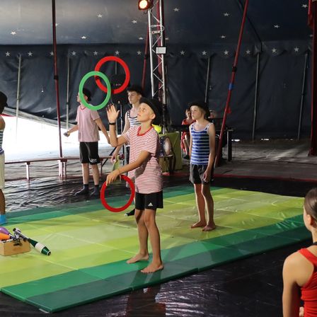 Eine Gruppe junger Kinder übt das Jonglieren bei einer Zirkusveranstaltung. Sie stehen auf einem bunten Teppich unter einem Zelt mit einem Sternenhimmel im Hintergrund.